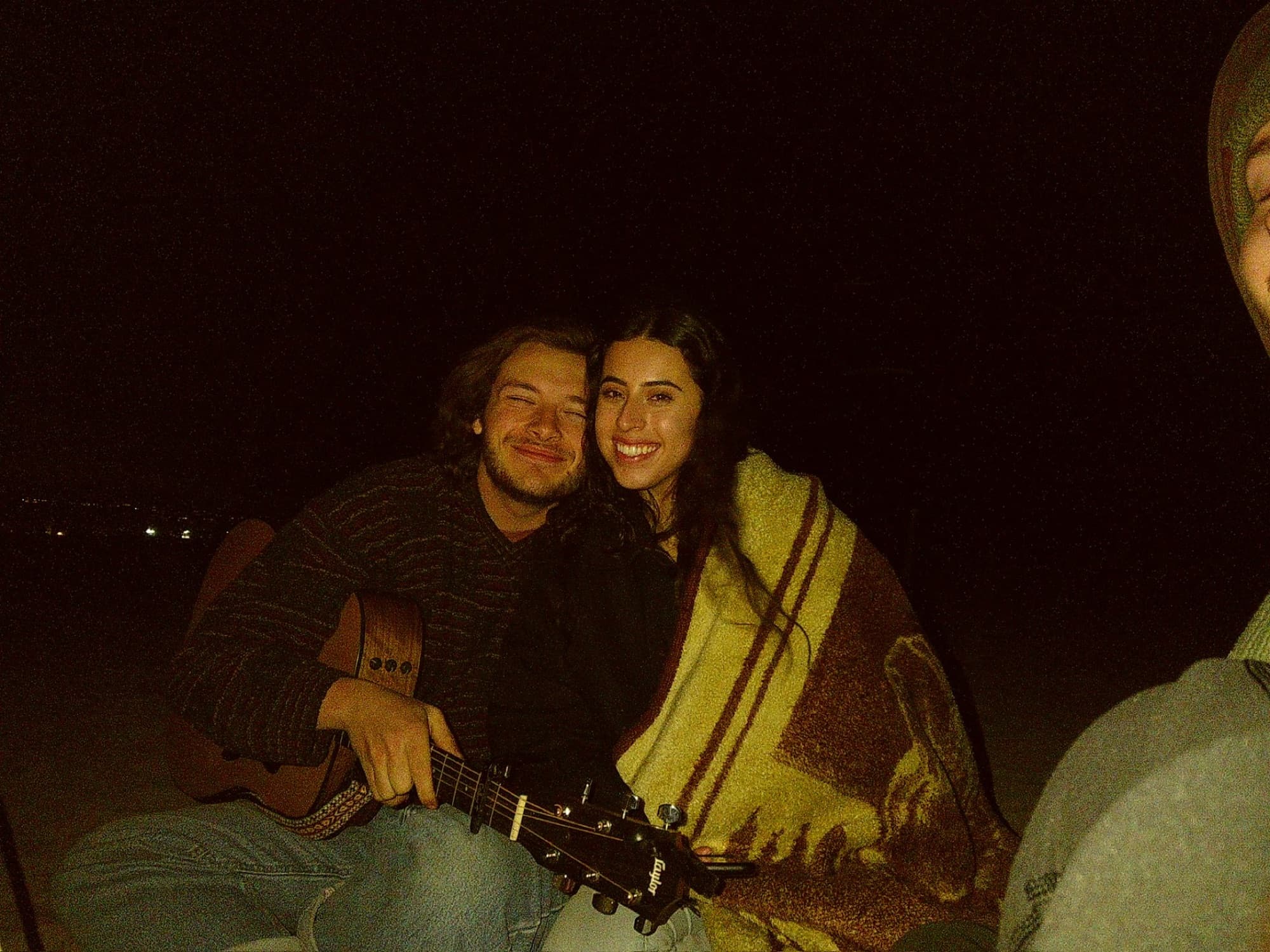 Colby and a friend wrapped in a blanket by a fire at night, an acoustic guitar across his lap